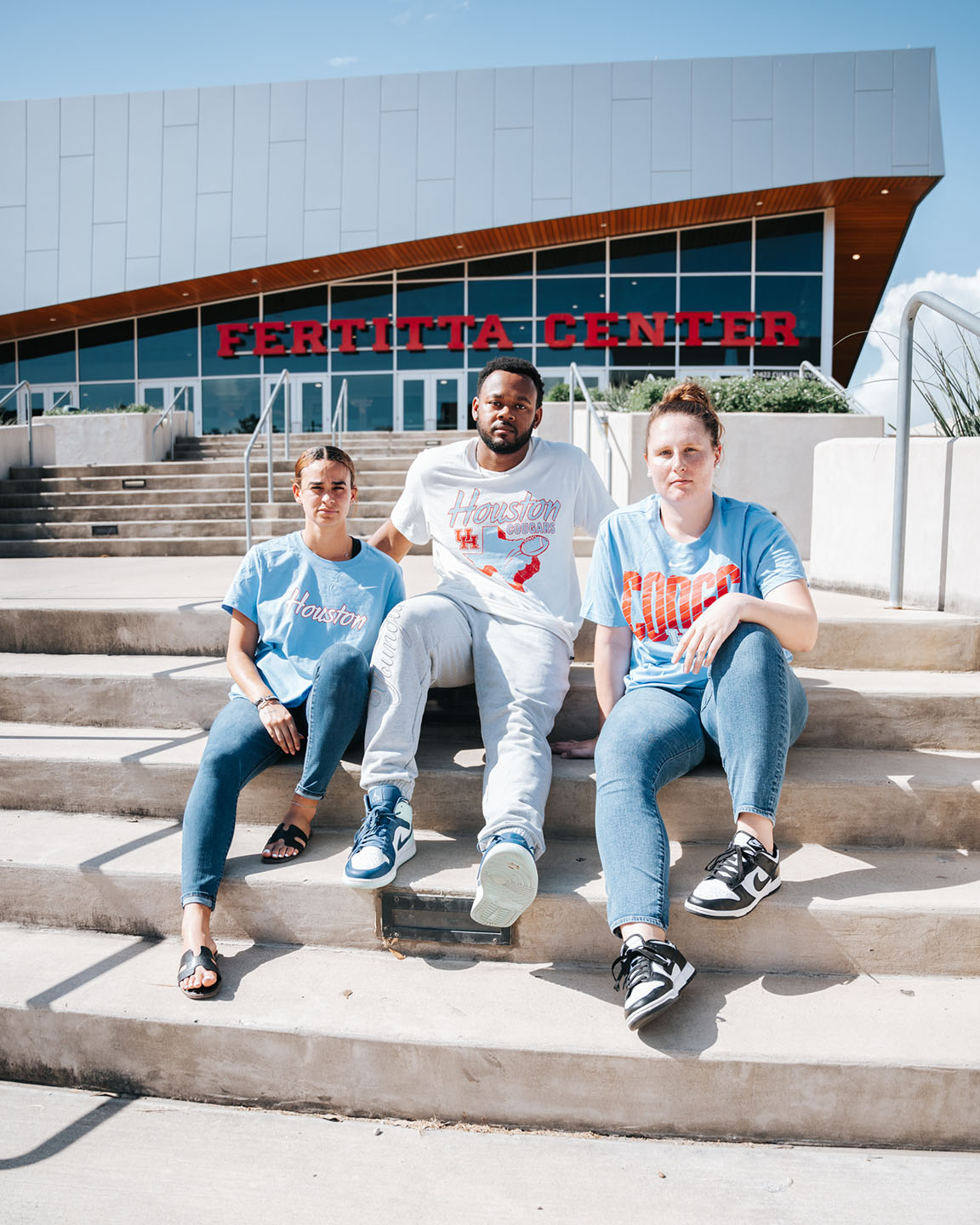 Three students sit together on the steps outside Fertitta Center while wearing Houston Blue merchandise.