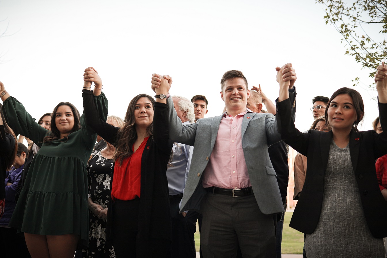 Students stand in a line holding each other's hands up while friends and family stand behind them