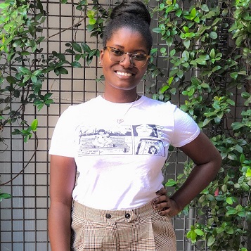 A woman with her black hair in a bun wearing glasses standing in front of a lattice panel with green vines