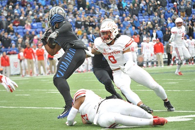 A football player in a white and red uniform reaches for a player in a black and blue uniform with a football in hand