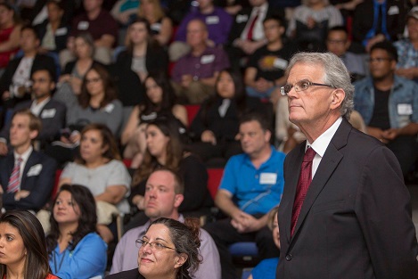 A silver-haired man in a suit with glasses standing next to a group of seated people