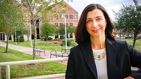 Portrait of a brown-haired woman in a dark suit with trees, a sidewalk and benches in the background