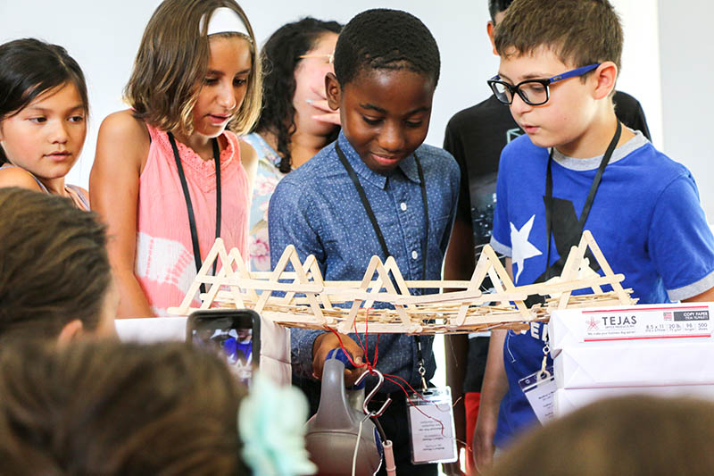 A young student hangs a weighted ball from a small suspension bridge made of craft sticks to test the structure. He is surrounded by his teammates and onlookers.