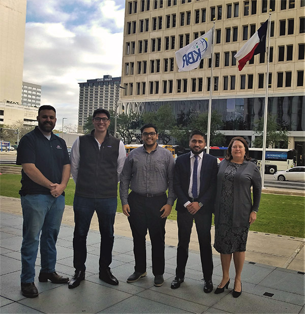 A group of four male students with a female instructor standing in from of a tall concrete building surrounded by other tall buildings