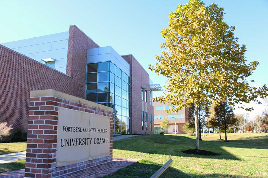 Photo of a large brick sign that says, “Fort Bend County Libraries. University Branch”. A large two story bricka nd glass building and a tall tree are behind the sign.